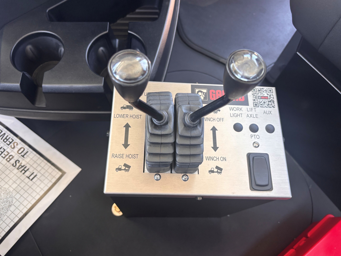 Joystick-style control panel with two levers labeled lower hoist and raise hoist, and worklift controls in a Mack GR64B cab interior.