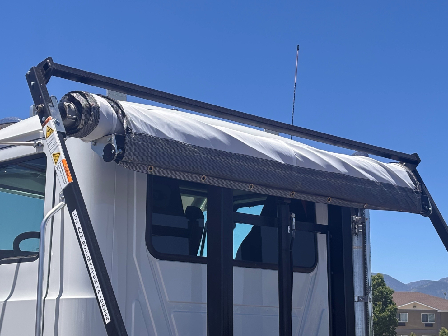 Mack GR64B roll-off truck cab side with folded tonneau and hydraulic roll-off system visible under bright blue sky.