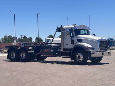Mack GR64B rolloff truck in side view with white cab and flatbed trailer, showing chassis, dual rear axles, and wheel components.
