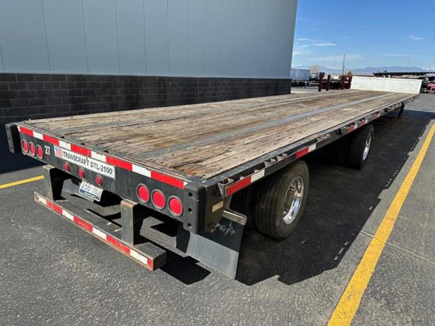 Flatbed trailer showing wood deck with rear lights, dual rear axles, and reflective tape along sides; back plate with Transcraft branding.