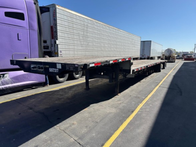Flatbed trailer in a truck yard with purple truck cab, long silver trailer, and other trailers in background.