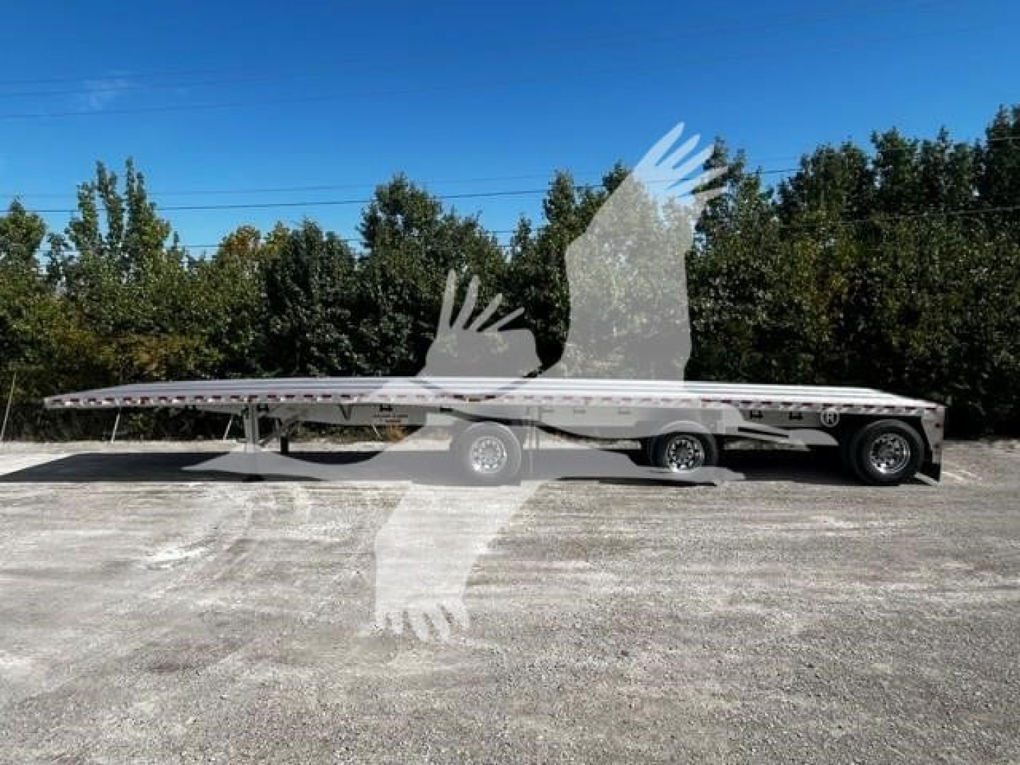 Flatbed trailer showing long deck with tandem axle at rear and single axle forward, VIN sticker visible, blue sky with trees in background.