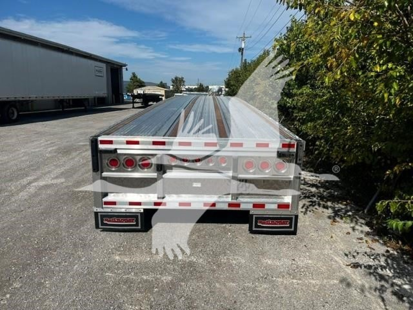Flatbed trailer rear showing taillights and mud flaps with brand plates, metal ramps folded on top, gravel lot, warehouse left, trees right.