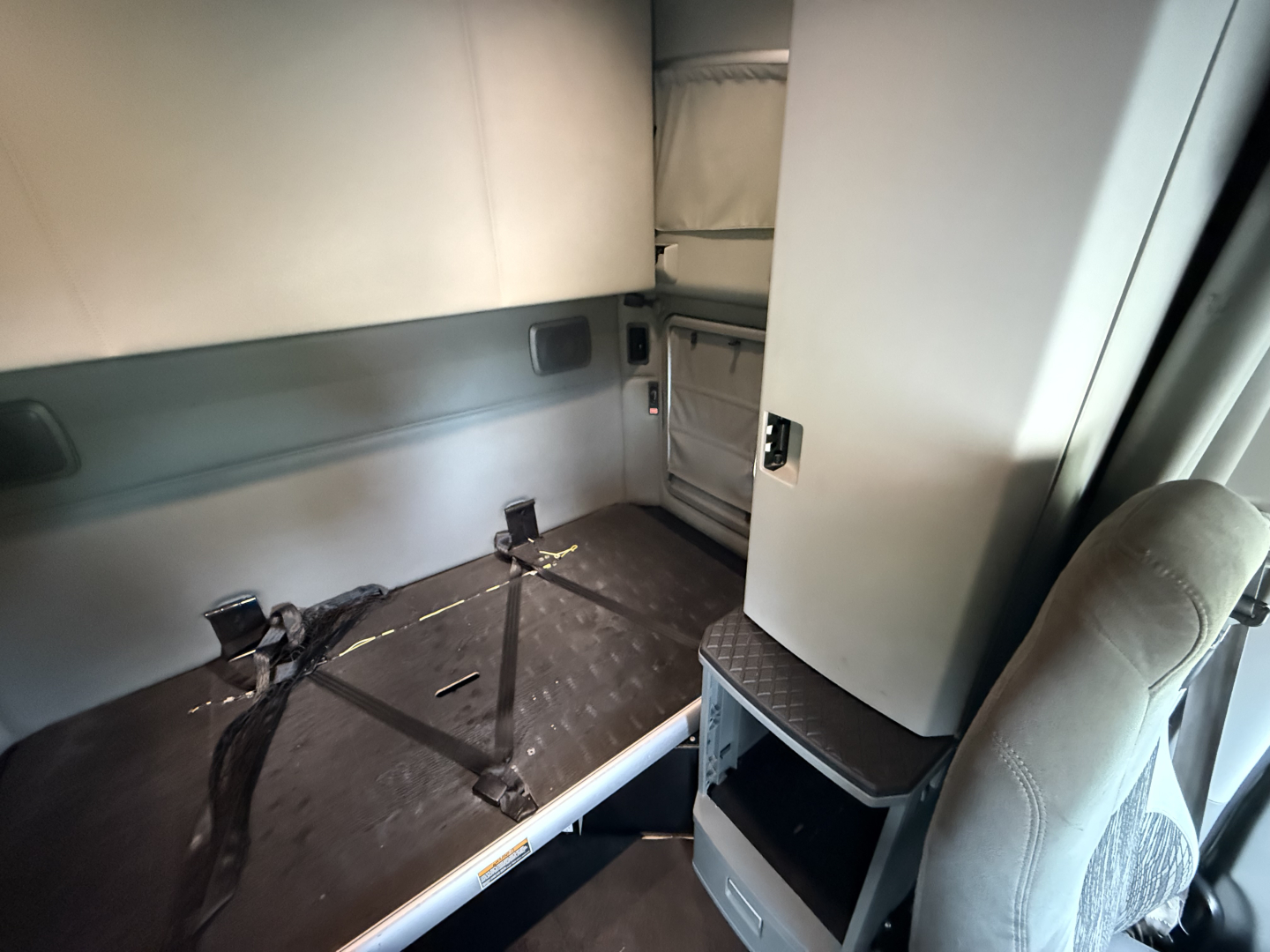 Truck cab interior of a 2021 Kenworth T680 sleeper showing bare floor with mounting brackets and side storage cabinet