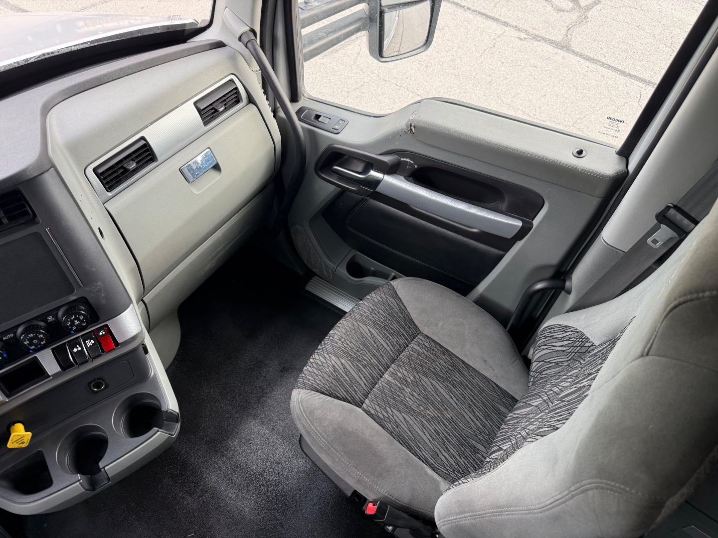 Kenworth T680 cab interior view showing driver's seat and dashboard with gray fabric seat, door panel, and cup holders.