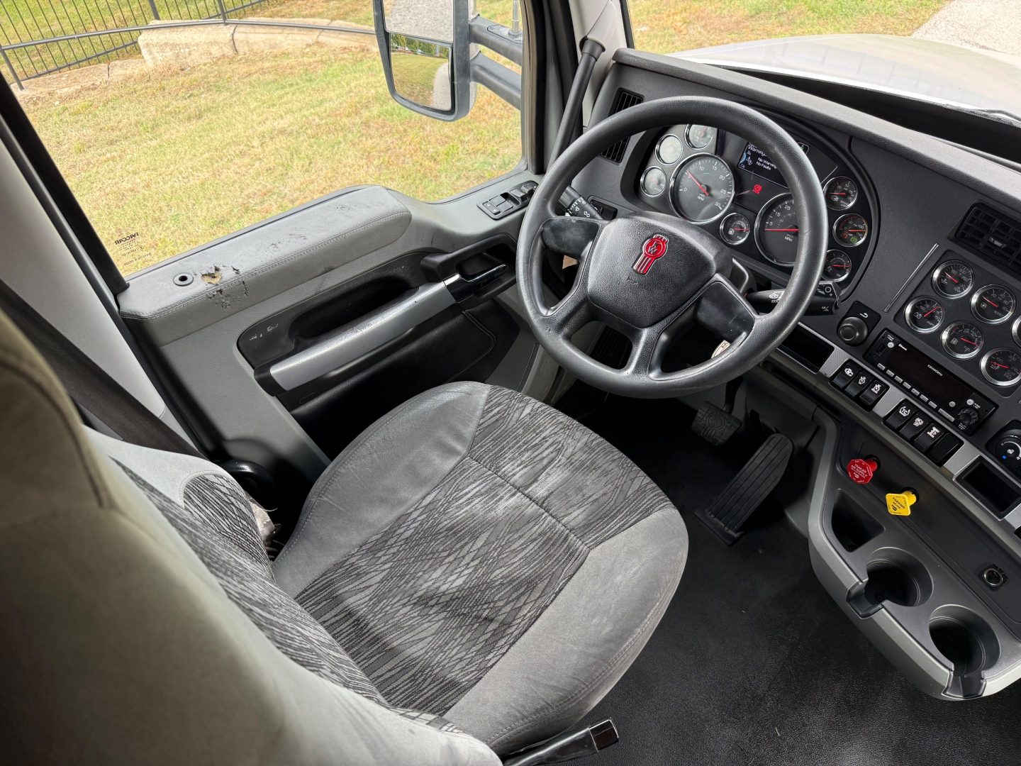 Kenworth T680 cab interior showing steering wheel with logo, dashboard gauges, and driver seat from the left door perspective.