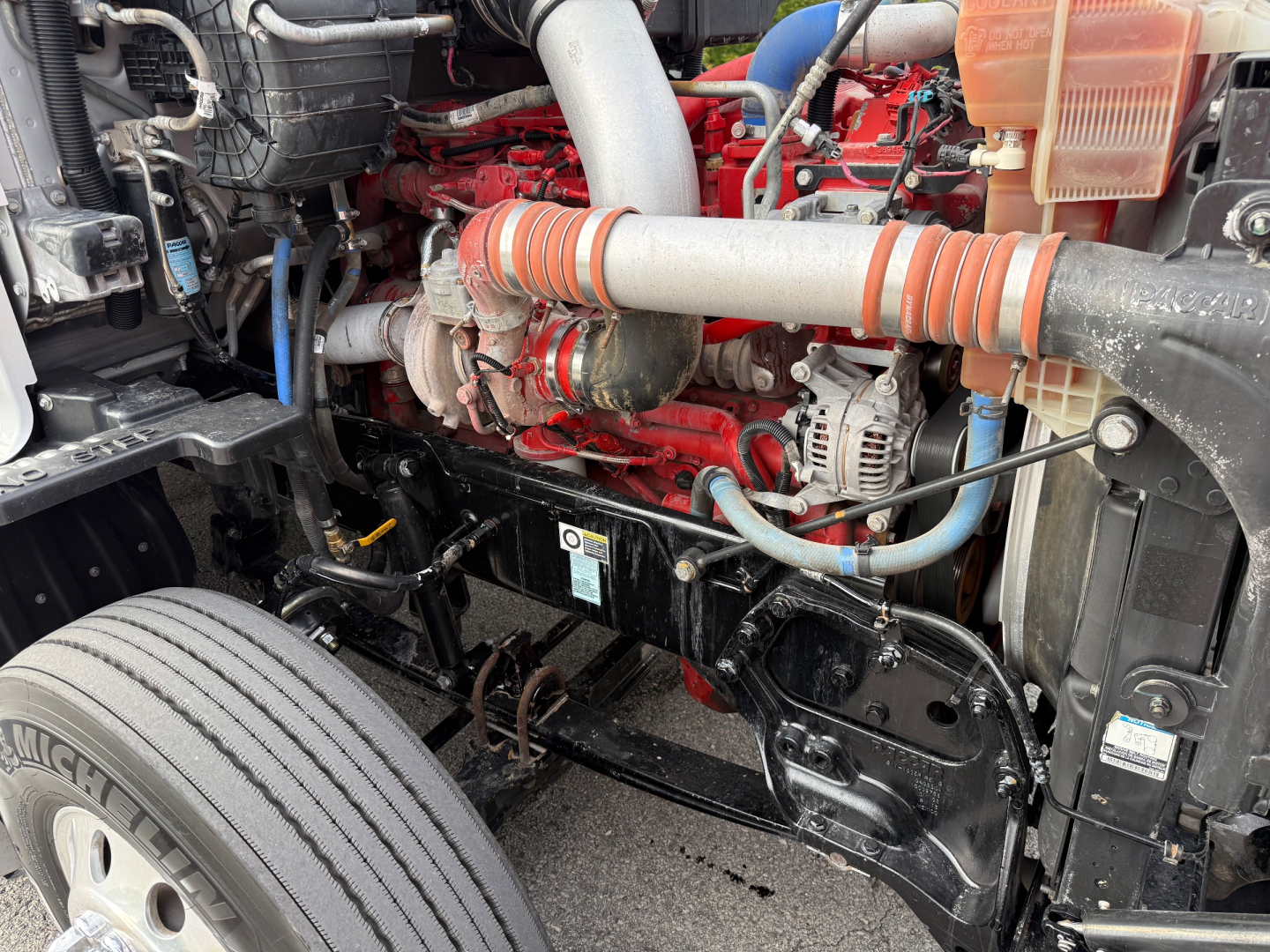 Kenworth T680 engine bay showing red diesel engine with orange striped intake hose, alternator, hydraulic lines, and front suspension area.