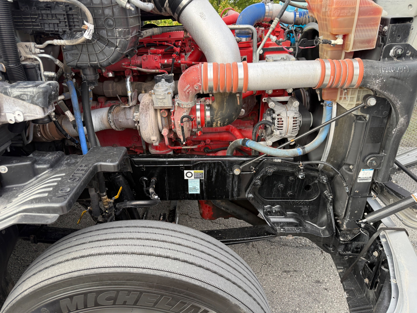 Kenworth T680 engine bay showing red engine block, turbo, hoses, alternator, and front suspension components.