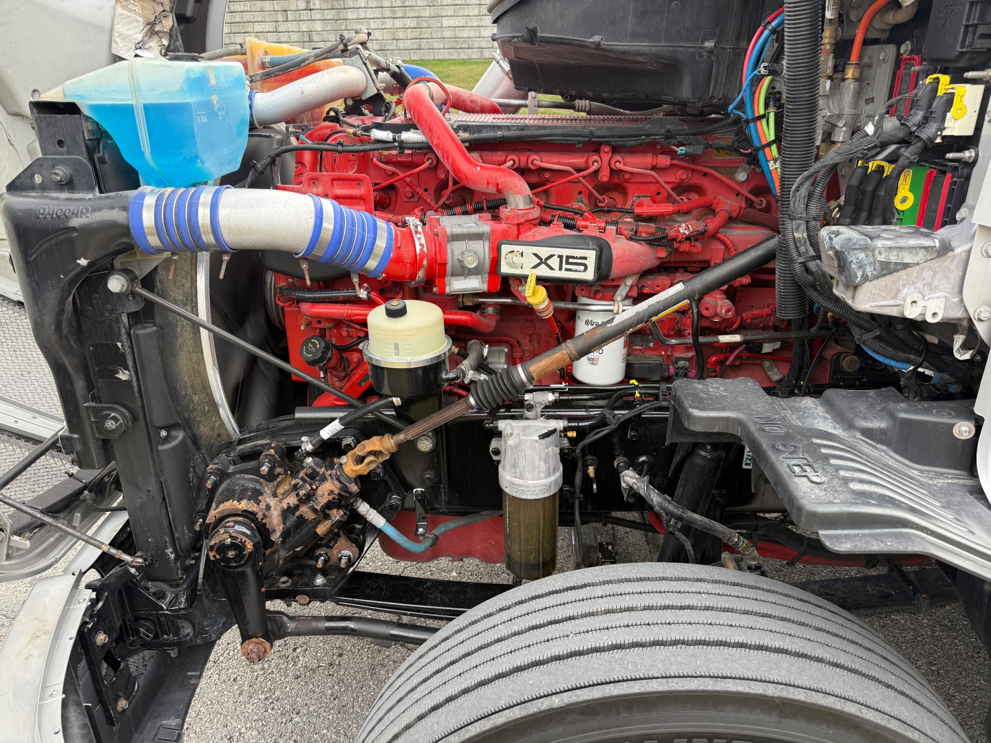 Kenworth T680 engine bay with red X15 engine, blue coolant reservoir, yellow cap, and various hoses and wiring visible.
