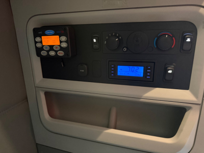 Cab interior dash of a Kenworth T680 showing control panel with blue digital radio display, round climate dials, a Carrier HVAC device, and a storage pocket.