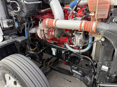 Kenworth T680 engine bay showing red diesel engine with orange striped intake hose, alternator, hydraulic lines, and front suspension area.