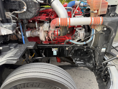 Kenworth T680 engine bay showing red engine block, turbo, hoses, alternator, and front suspension components.