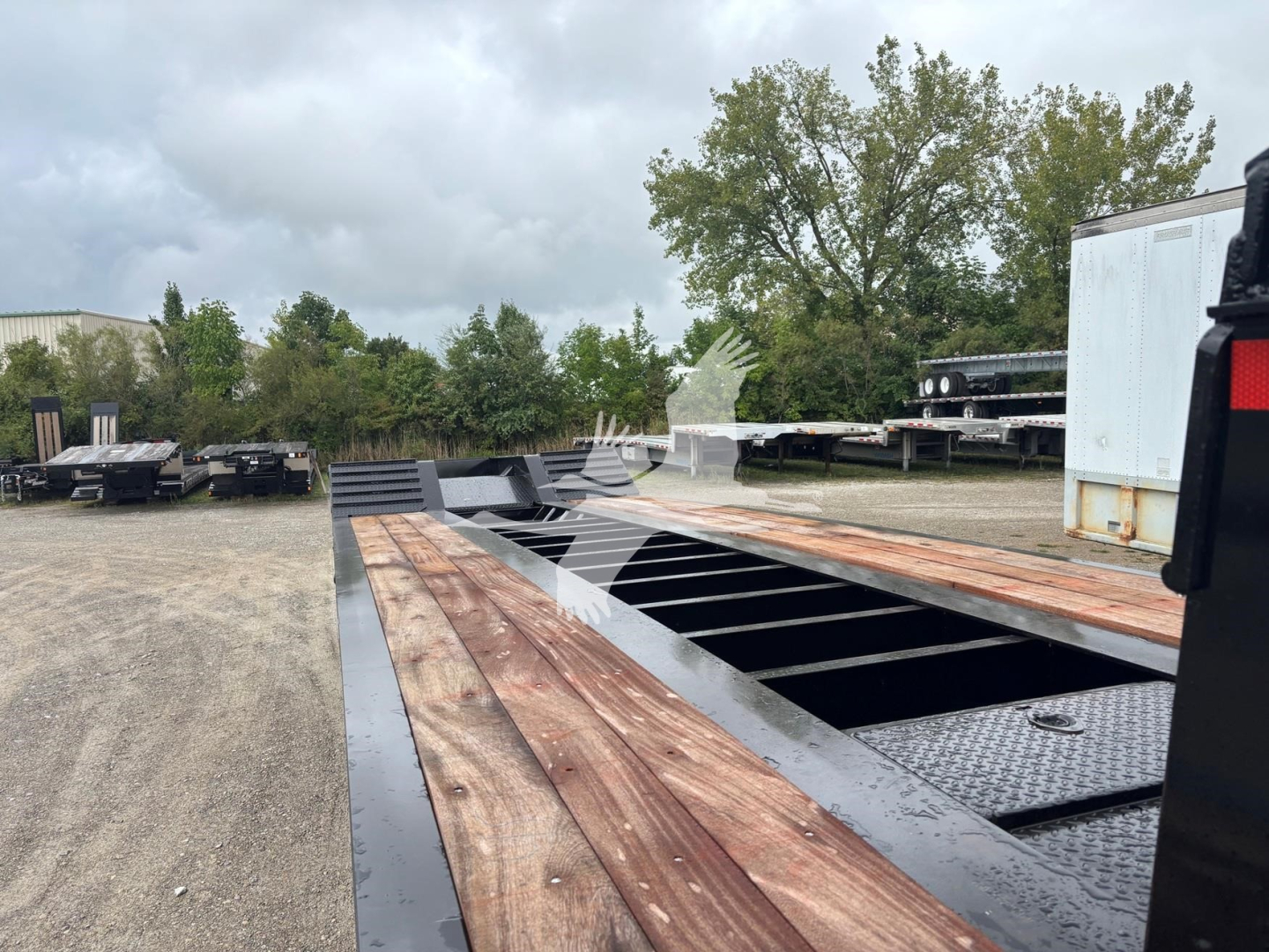 Lowboy trailer bed with wooden deck planks and metal frame, daytime outdoor lot showing long flatbed sections and a few trailers in the background.