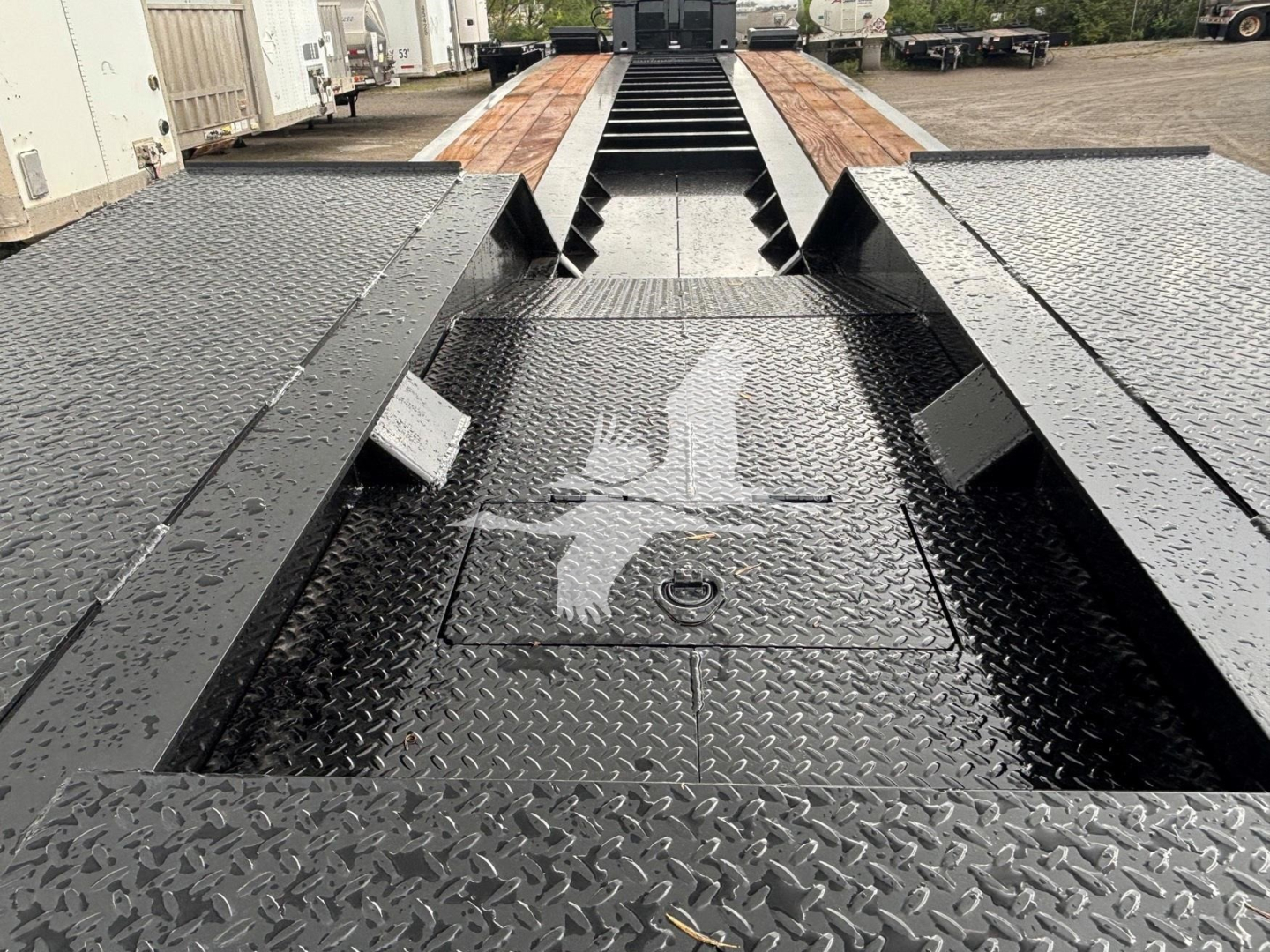 Lowboy trailer deck with textured black steel, central spine and cutouts; raised gooseneck slots visible, wet surface with diamond plate pattern.