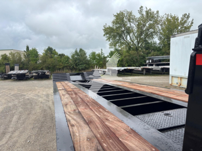 Lowboy trailer bed with wooden deck planks and metal frame, daytime outdoor lot showing long flatbed sections and a few trailers in the background.
