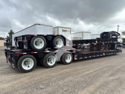 Flatbed trailer with multiple axles carrying a small metallic equipment container and spare tires on a rural lot.