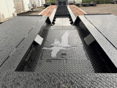 Lowboy trailer deck with textured black steel, central spine and cutouts; raised gooseneck slots visible, wet surface with diamond plate pattern.