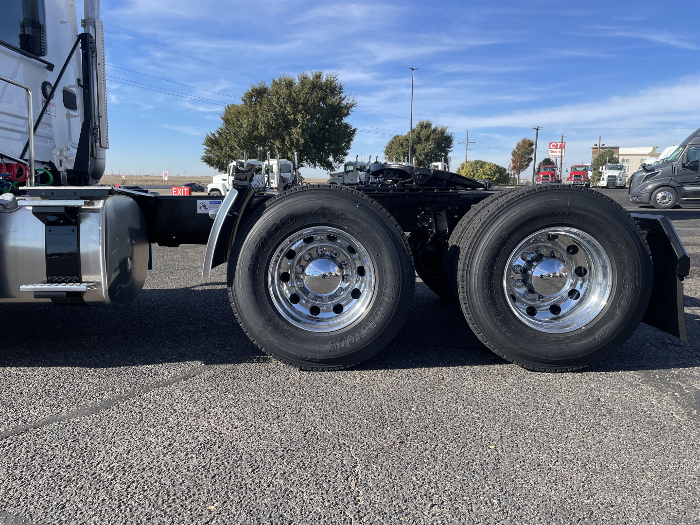 Dual rear axles of a Mack PR64T truck chassis showing chrome rims and tires, with cab visible on left and fifth wheel mount area; clear blue sky and dealership lot in background.