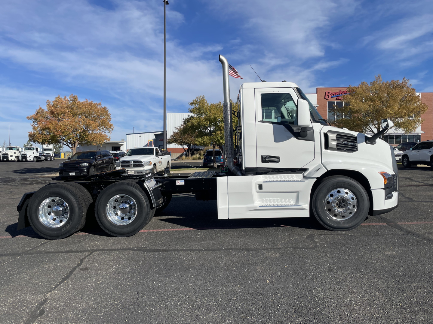 Truck chassis side profile of a white Mack PR64T tractor in a parking lot, showing cab, low air intake, and dual rear axles with chrome wheels.
