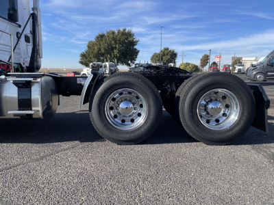 Dual rear axles of a Mack PR64T truck chassis showing chrome rims and tires, with cab visible on left and fifth wheel mount area; clear blue sky and dealership lot in background.