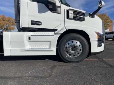Truck side view showing front wheel and lower cab detailing on a white 2026 Mack PR64T with chrome wheel hub, steps, and door handle.
