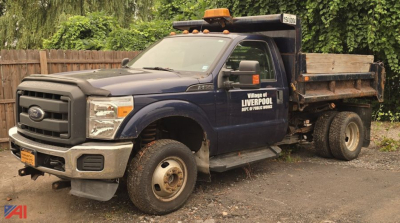 Ford F-350 Super Duty XL dump truck with V-plow, side text shows Village of Liverpool, NY, dual rear wheels, orange beacon on cab, dirt/grime on body.