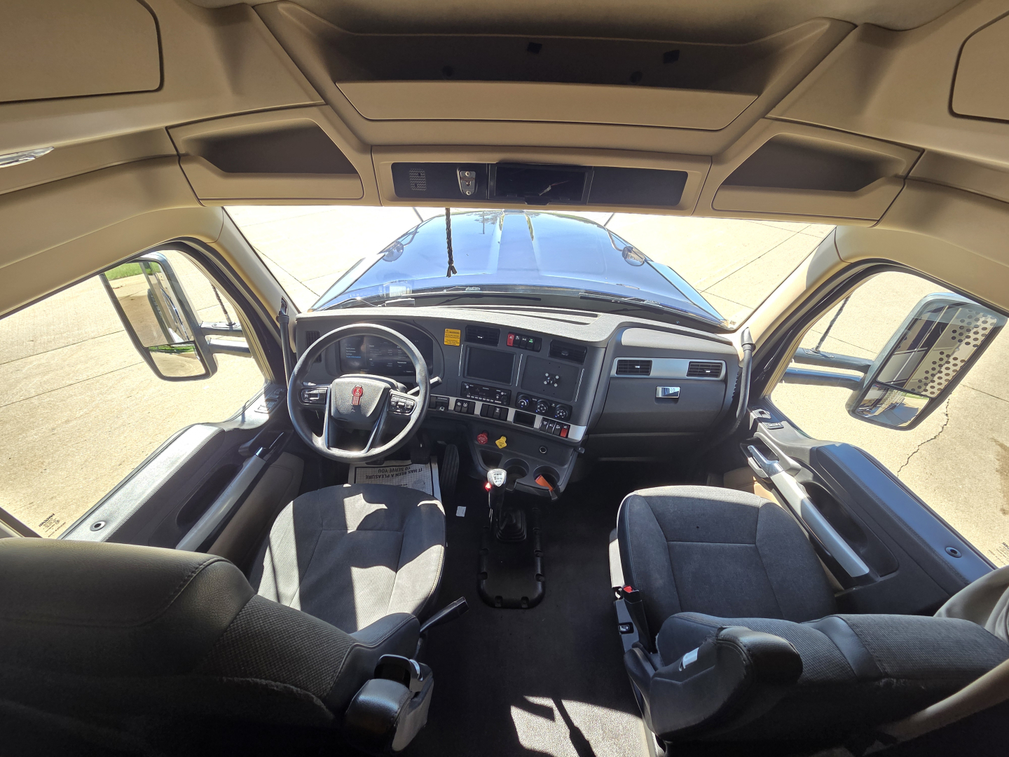 Kennworth W990 cab interior view from front windshield showing steering wheel, dashboard, and two seats with light gray upholstery.