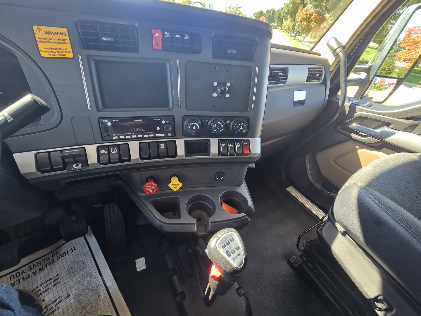 Kenworth W990 cockpit interior showing dashboard with multi-function display screens, control switches, steering column, gear shifter, and driver seat. Visible labels include warning stickers and dashboard controls.