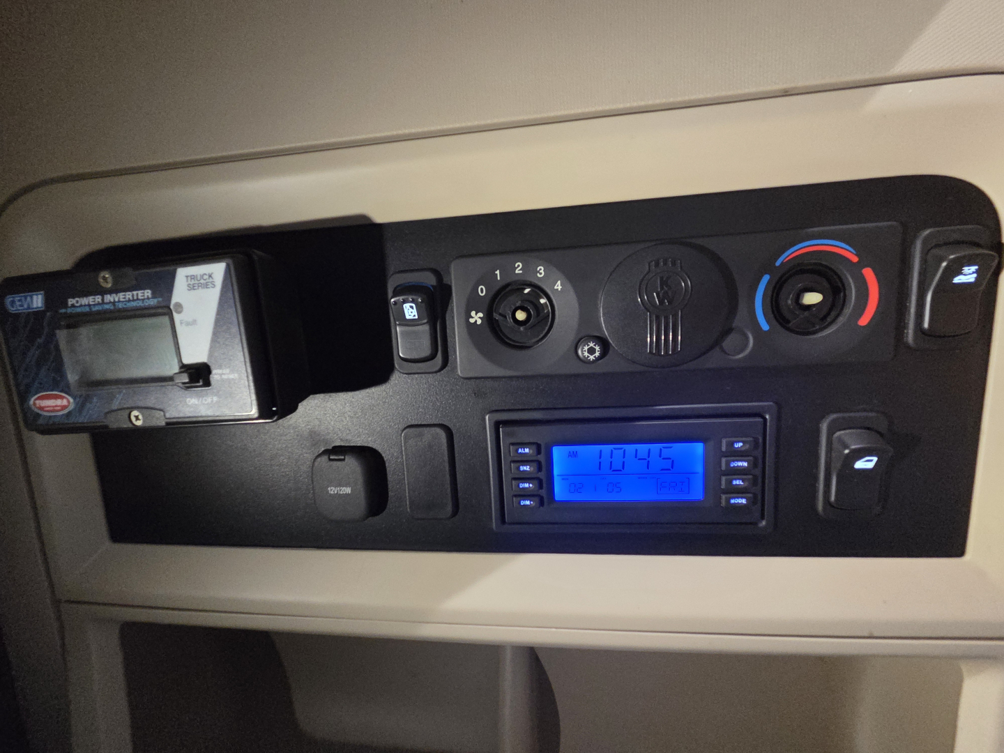 Kenworth W990 cab interior dash showing control panel with digital display and gauges. Visible text includes 'Power Inverter' on left device and blue backlit display; circular knobs and climate controls; no clear VIN or numbers readable.
