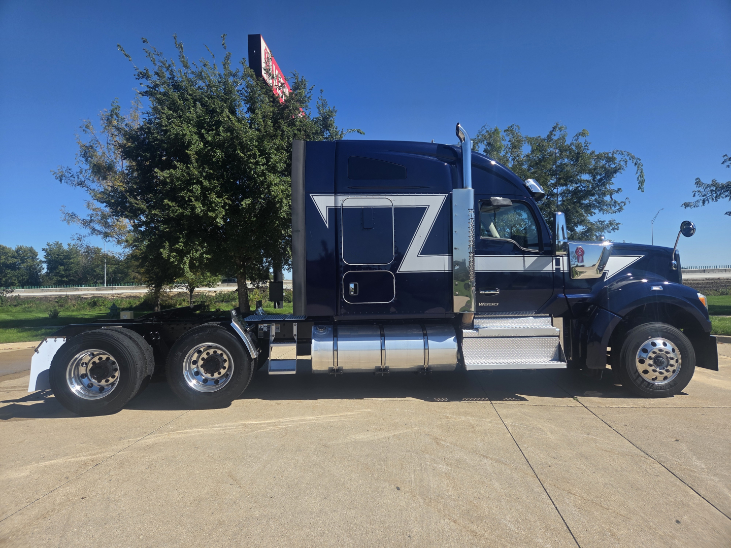Kenworth W990 conventional sleeper truck side view showing large Z-shaped graphic on dark blue cab, chrome fuel tanks, and six wheels at rear.