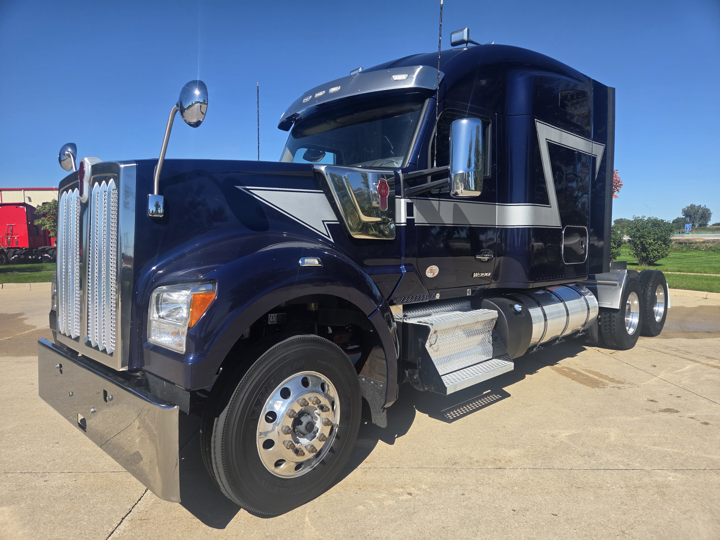 Kenworth W990 conventional sleeper truck in dark blue showing chrome grille, dual fuel tanks, side steps, and large front bumper.