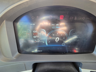 Dashboard display showing digital gauges in a Kenworth W990; center dial shows speed 0 MPH, various icons and white text; left and right indicators with numbers; below the speedometer is a small odometer reading; top edge shows glare and in