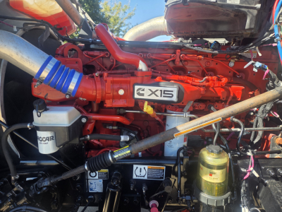Kenworth W990 engine bay showing Cummins X15 engine cover and red-painted components with hoses, reservoir bottles, and wiring in view.