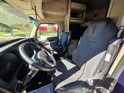Kenworth W990 truck interior showing driver seat, steering wheel, and front cabin area with side window and overhead storage.