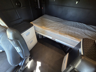 Inside Kenworth W990 sleeper cab showing a raised bed with mattress, storage drawers, and seat; visible panels and cabinetry.