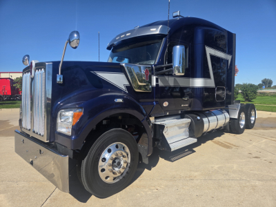 Kenworth W990 conventional sleeper truck in dark blue showing chrome grille, dual fuel tanks, side steps, and large front bumper.