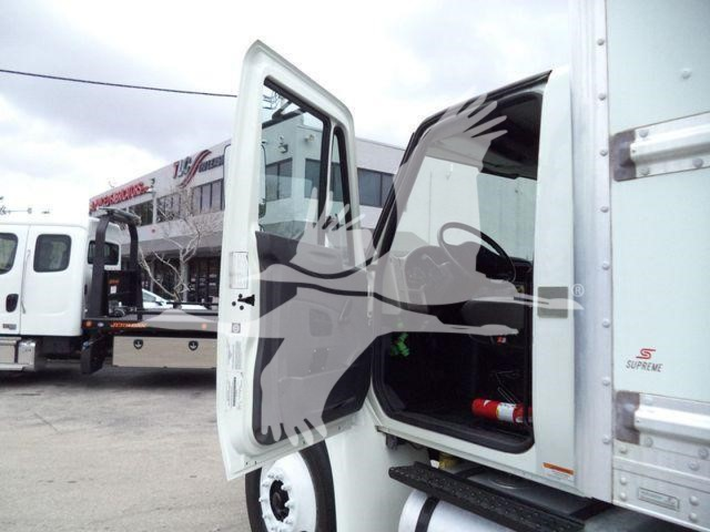 Truck cab interior with driver door open showing storage area and steps; white box truck side; other trucks in background.