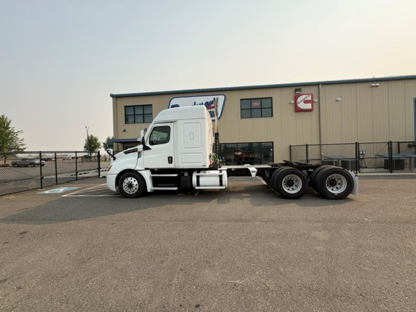Truck side view of a 2020 Freightliner Cascadia 126 sleeper tractor chassis in a lot. White cab, three rear axles, B3832 stock number visible, building in background.