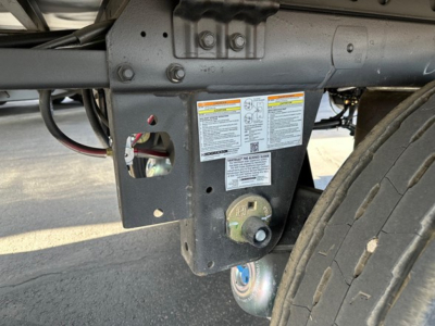 Utility trailer hitch area showing frame with warning labels and wheel, 2026 Utility 3000R trailer. Visible stickers with safety instructions and a coupling/kingpin nearby.
