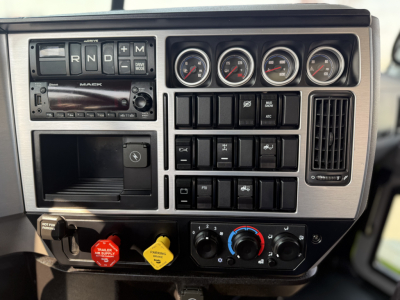 Mack AN64T truck dashboard showing gear selector, four round gauges, climate controls, and various switches with labeled red and yellow knobs.