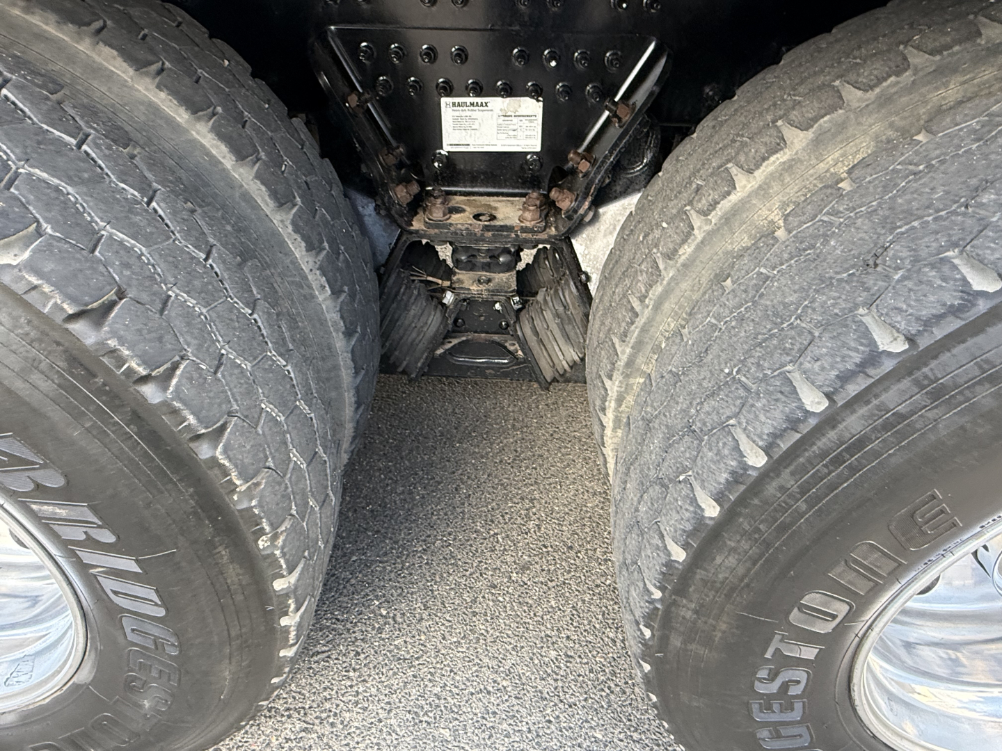 Rear view undercarriage showing dual rear wheels and drivetrain components of a Mack GR64B truck, with label placard mounted on frame between axles.