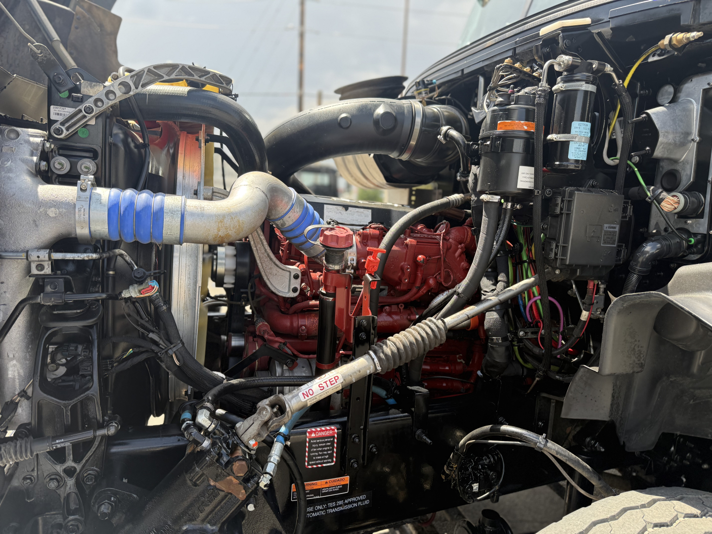 Mack GR64B engine bay showing red engine block, blue silicone intercooler hose, various black hoses, electrical wiring, and metal components.