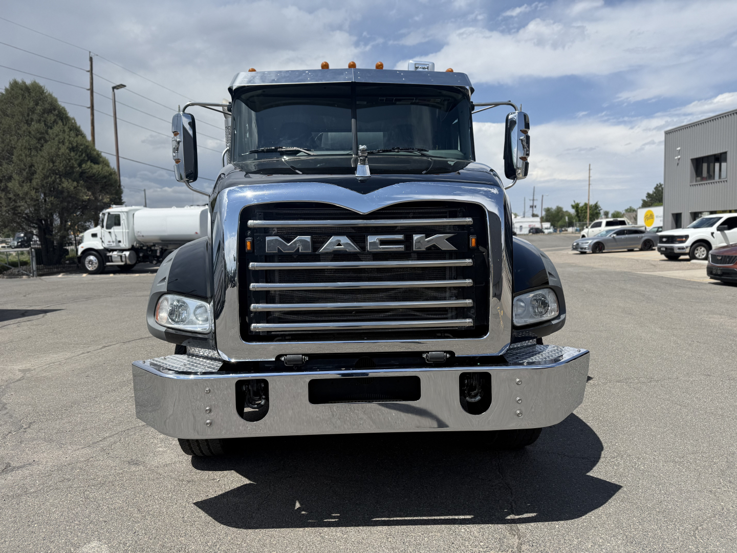 Mack GR64B heavy truck front view showing chrome grille with 'MACK' emblem and large bumper. Clear sky, dealership lot in background.
