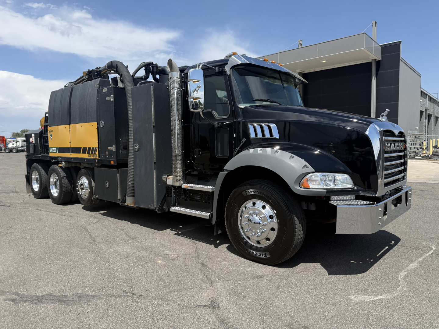 Vehicle side view of a 2023 Mack GR64B vacuum truck with black cab and yellow-accented vacuum tank, dual rear axles, and chrome bumper.