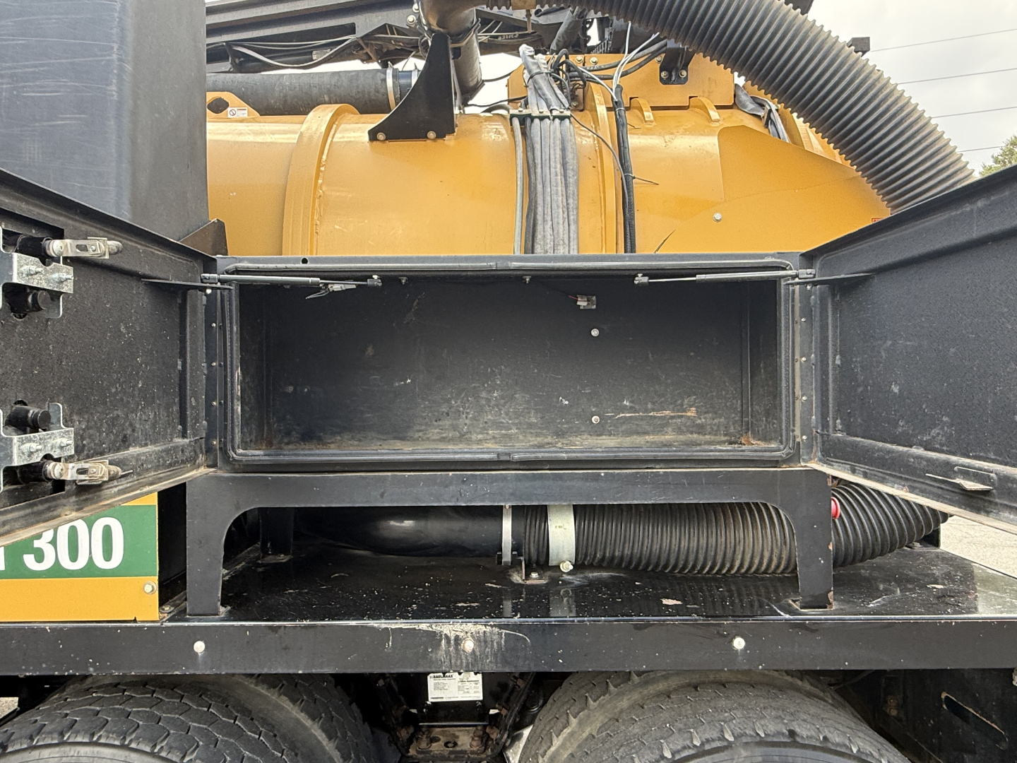 Truck chassis showing empty storage compartment and vacuum hose with yellow machinery in background; visible tires and metal frame.