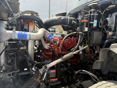 Mack GR64B engine bay showing red engine block, blue silicone intercooler hose, various black hoses, electrical wiring, and metal components.