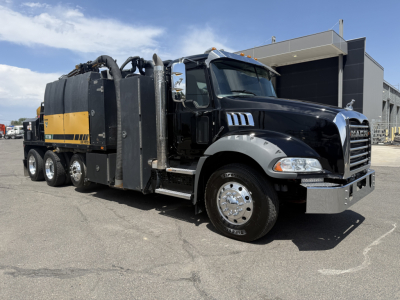 Vehicle side view of a 2023 Mack GR64B vacuum truck with black cab and yellow-accented vacuum tank, dual rear axles, and chrome bumper.