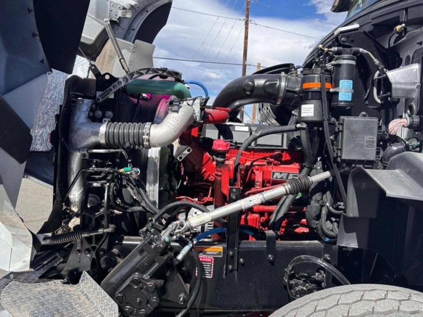 Tractor-trailer truck engine bay view showing red Mack engine with hoses, wiring, and components; labels and fittings visible.