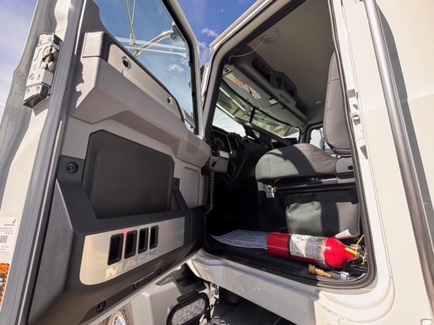 Truck cab interior view from open door showing dashboard, steering wheel, and passenger seat in a Mack GR64B roll-off truck.