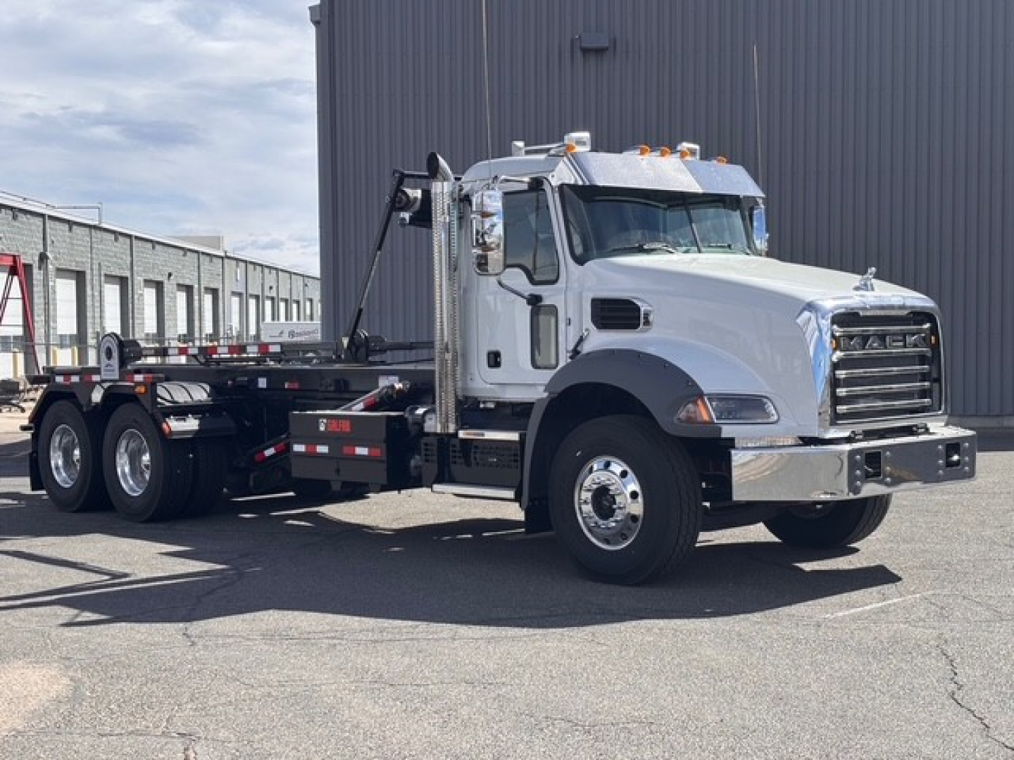 2026 Mack GR64B roll-off truck in white with a black hook-lift system, chrome grille, and dual rear axles. Side text includes Mack logo on grille and reflective strips.